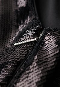 Chaqueta negra de lentejuelas con un tirador de cremallera plateado con "SUPERDRY" grabado. Primer plano que muestra la intrincada textura y diseño de las lentejuelas.