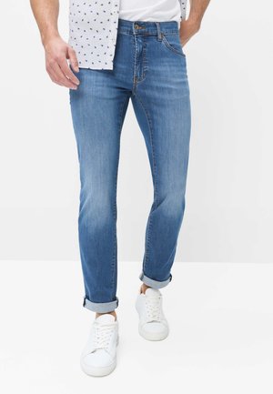 Mann trägt hellblaue Slim-Fit-Jeans mit aufgerollten Bündchen, weiße Sneaker, teilweise sichtbares weißes Hemd, steht vor einem schlichten weißen Hintergrund.