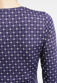 Tmavě modrý top s dlouhými rukávy a diamantovým vzorem, zdobený malými bílými květinovými akcenty a strukturovaným lemováním kolem výstřihu.