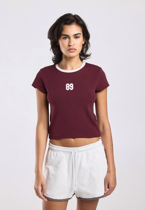 Jonge vrouw die een wijnrood cropped t-shirt draagt met witte '89' aan de voorkant en witte sportshorts met trekkoord tegen een effen achtergrond.