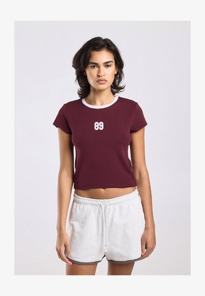 Jonge vrouw die een wijnrood cropped t-shirt draagt met witte '89' aan de voorkant en witte sportshorts met trekkoord tegen een effen achtergrond.
