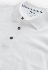Hvid polo shirt lavet af tekstureret stof, med en tre-knaps lukning, sorte knapper, og en krave med røde og blå detaljer.