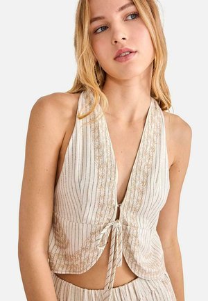 Mujer joven con cabello rubio que lleva una blusa sin mangas color beige con rayas verticales y detalles bordados, atada al frente.