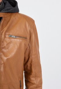 Brun læderjakke med en lynlåslukket brystlomme, glat tekstur og en afslappet hætte lavet af gråt stof.