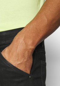 En persons hand delvis inne i framfickan på svarta byxor, med en ljusgul skjorta synlig ovanför midjebandet.