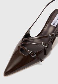 Scarpa con tacco alto in pelle verniciata marrone, con punta affusolata, caratterizzata da cinghie incrociate e fibbie in tonalità argento. Finitura liscia e lucida.
