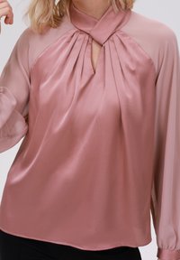 Blouse en satin rose clair avec un décolleté en forme de clef, un devant plissé et de longues manches transparentes. Texture lisse et détails drapés élégants.