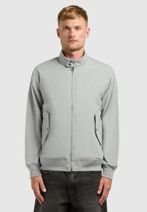 Man with short hair wearing a light gray zip-up jacket with buttoned flap pockets and dark jeans, facing forward against a plain background.