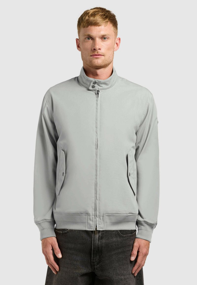 Homme aux cheveux courts portant une veste zippée gris clair avec des poches à rabat boutonnées et un jean foncé, faisant face à l'avant sur un fond uni.