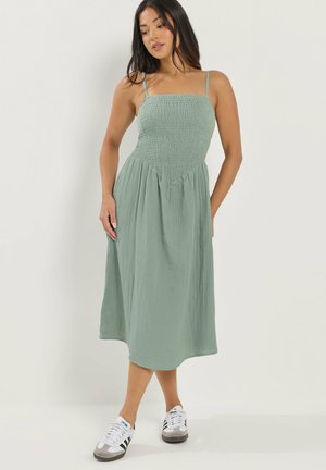 Kobieta ubrana w zieloną, jasnozieloną, midi sukienkę bez rękawów z marszczonym gorsetem i białe trampki, stojąca na jednolitym jasnym tle.