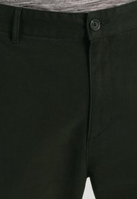Ciemnozielone spodnie z mocnej tkaniny, z zapięciem na guzik, przednimi kieszeniami i gładką fakturą, z widocznymi detalami szwów.