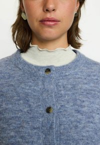 Mėlynas mezginys kardiganas su apvaliu iškirpimu, su sagų užsegimu ir tekstūruota medžiaga. Dėvimas ant balto, bangų formos apykaklės topai.