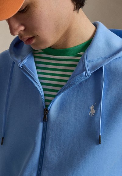 Noor inimene kannab oranži mütsi, rohevalgete triibulist särki ja hele sinist tõmblukuga kapuutsi valge polo mängija logoga.