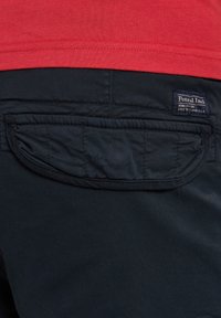 Marinebukser med en quiltet lomme og et syet logo mærke, parret med en rød t-shirt, der ligger ovenpå. Stoffet fremstår glat og holdbart.