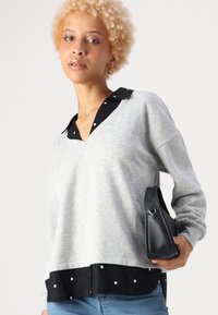 Sweatshirt gris avec un col et un ourlet en pois noirs. Manches longues, coupe décontractée et design superposé. Porte un petit sac à main noir.