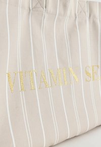 Béžová látková taška s bílými vertikálními pruhy a zlatým textem částečně znějícím „VITAMIN SEA“.