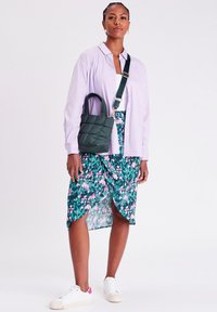 Borsa verde gonfiata con due manici e una tracolla, indossata con una camicia button-up viola chiaro e una gonna verde a fantasia floreale. Sneakers bianche.