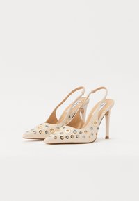 Beige slingback high heels with pointed toe, adorned with metallic grommets, and a thin stiletto heel. Smooth texture provides a sleek finish.