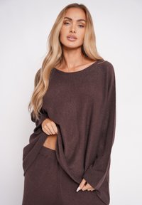 Brązowy sweter dzianinowy o luźnym kroju, szerokim dekolcie i długich rękawach, zestawiony z pasującymi prążkowanymi spodniami. Miękka tekstura, minimalne wykończenie.