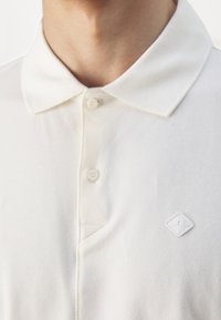 Vit polo skjorta med klassisk krage, tre knappar och en liten diamantlogotyp på bröstet. Tyget verkar vara texturerat.