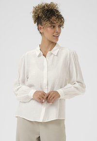 Žena s kudrnatými vlasy v bílé dlouhé košili na knoflíky a béžových kalhotách, dívající se do strany na pozadí s jemným světlým tónem.