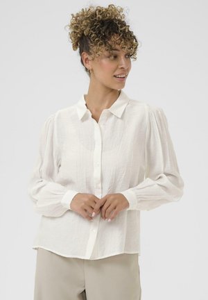 Frau mit lockigem Haar in einer weißen Bluse mit langen Ärmeln und beigen Hosen, die zur Seite blickt, vor einem schlichten hellen Hintergrund.
