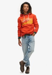 Rød hættetrøje med bjerggrafik, hvide snøringer, foran kængurulomme. Matchet med lyseblå jeans og brune støvler. Tekstureret bomuldsblanding.