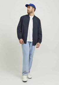 Tamsiai mėlynas bombonešio švarkas su priekinės užtrauktuku, balta marškinėliai, šviesiai mėlyni džinsai ir balti sportbačiai. Modelis dėvi mėlynę kepurę.