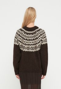 Brown knit sweater featuring a fair isle pattern in cream, with a round neckline and long sleeves, worn over a pleated brown skirt.