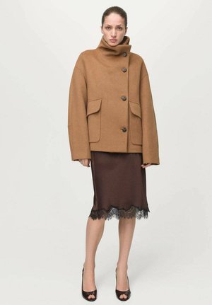 Abrigo de lana marrón con corte extragrande, cuello alto y grandes bolsillos frontales, combinado con una falda de seda marrón con ribete de encaje negro.
