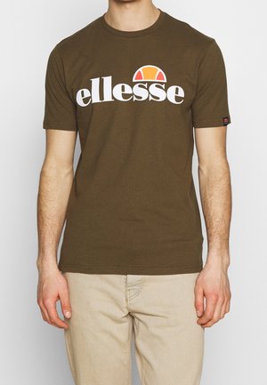 Mand iført en olivengrøn ellesse t-shirt med hvid og orange logo, parret med lysebeige bukser, stående mod en ensfarvet baggrund.