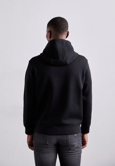 Svart huvtröja med ribbade mudd och fåll, med en mjuk textur och minimalistisk design. Bärs med grå jeans, sedd bakifrån.