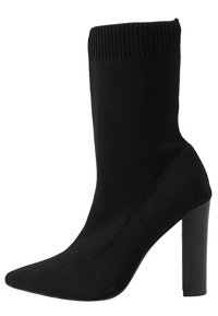 Czarna skarpetka na obcasie z dzianiny, z wyraźnym noskiem i wysokim słupkowym obcasem. Górna część ma ribbowane mankiety, co dodaje tekstury i lepszego dopasowania.