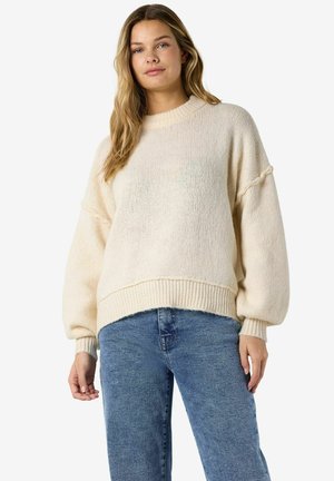 Jeune femme aux longs cheveux châtain clair portant un pull en grosse maille crème et un jean bleu, debout devant un fond blanc.