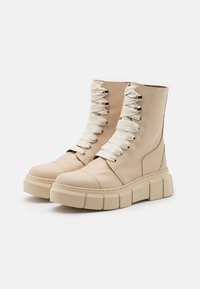 Botas de tornozelo em pele bege com solas de plataforma grossas, bicos redondos e atacadores planos grandes. Design simples com ilhós metálicos e detalhes costurados.