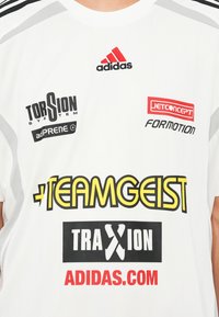 Bílé sportovní dres s černými a šedými akcenty, obsahující více log v černé, červené a žluté barvě. Texturovaná tkanina s vertikálními pruhy.