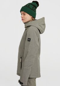 Grijze geïsoleerde jas met een gestructureerd patroon, verstelbare capuchon en ritsen, gecombineerd met een groene gebreide muts met zwart lettertype en een pompon.