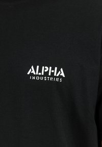 Μαύρο ύφασμα με λευκό κείμενο "ALPHA INDUSTRIES" τυπωμένο με γράμματα σε στένσιλ στο σημείο του στήθους ενός ρούχου.