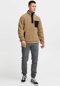 Sudadera de forro polar color crema con media cremallera, bolsillo negro en el pecho y bolsillos laterales. Pantalones grises y zapatos negros completan el atuendo. Textura suave.