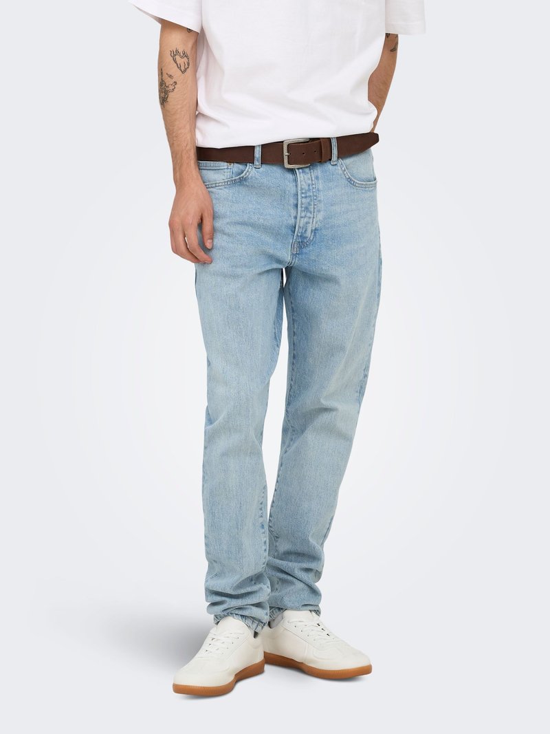 Man draagt lichtblauwe jeans, witte sneakers met rubberen zolen, bruine riem en wit T-shirt, staand tegen een effen achtergrond.