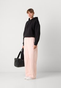 Sweatshirt à capuche noir, sweatpants rose clair et baskets blanches. Tenant un sac de sport noir. Matériaux doux avec une coupe décontractée.