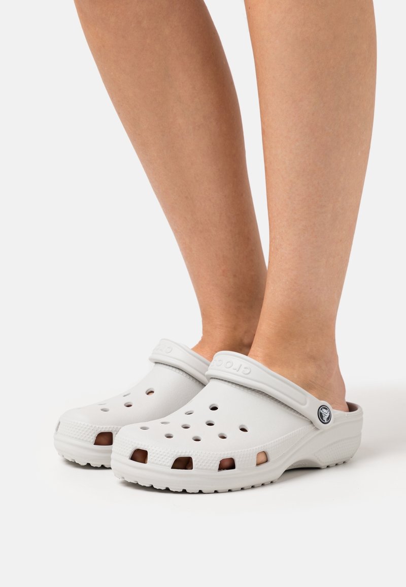 Sabots gris clair à enfiler avec des trous de ventilation portés aux pieds sur un fond blanc uni.