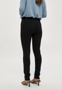 Zwarte smalle broek van rekbaar materiaal met een gladde textuur, slank ontwerp en discreete achterzakken, gecombineerd met grijze haksandalen.
