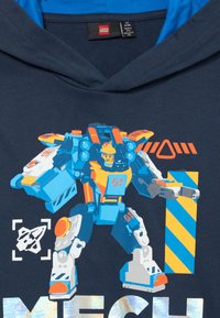 Lego robots ar smaidīgu figūru iekšpusē, zilos un oranžos toņos, stāv blakus svītrainai zilu un dzeltenu bloku uz tumši zilas auduma apkakles fona.
