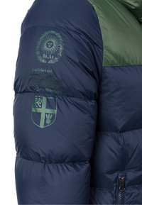 Marinblå puffjacka med gröna accenter, prydd med broderade logotyper och en dragkedja. Ytan ser jämn och isolerad ut.