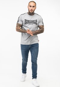 Gray t-shirt with black "Lonsdale London" logo, short sleeves, and black side stripes, paired with blue jeans and white sneakers.