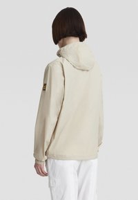 Giacca a vento beige con cappuccio, polsini elasticizzati e un'etichetta con logo sulla manica sinistra. Texture liscia, design leggero, adatta per essere indossata a strati.