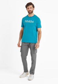 Teal cotton t-shirt with "TOG24" graphic, paired with grey cargo pants featuring side pockets. Model wears white sneakers.