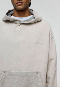 Lichtbeige sweatshirt met een grijze gestructureerde capuchon, voorzak en subtiele borduursels. Kenmerkt zich door metalen drukknoopsluitingen en zachte stof.