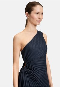 Asymetryczna granatowa sukienka charakteryzuje się teksturowanym materiałem, plisowanym wzorem oraz wycięciem w talii. Sparkle akcenty dodają subtelnego blasku.